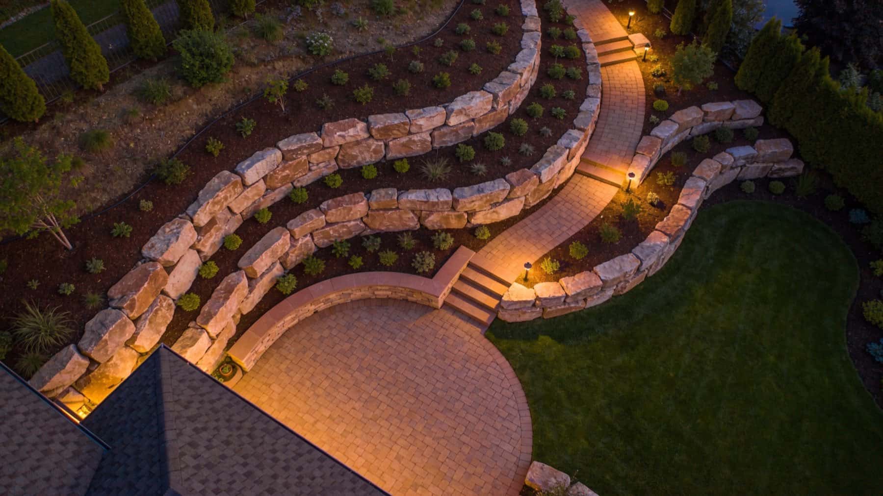 Aerial view at dusk of terraced boulder retaining walls stepping down a slope, with a lit paver path, steps and a round paver patio beside a lawn